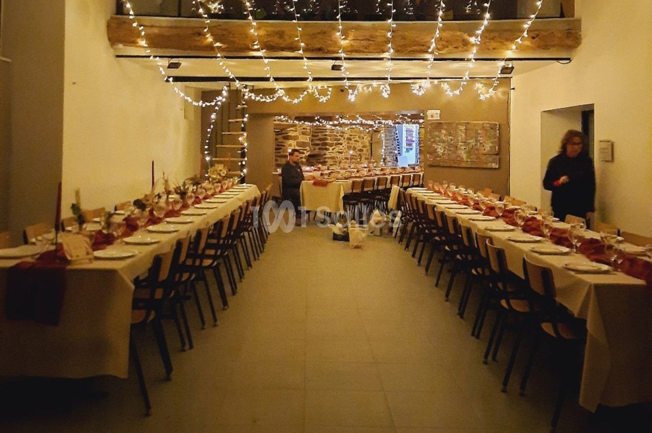 Salle décorée pour un repas festif, avec longues tables dressées, guirlandes lumineuses suspendues et murs en pierre.