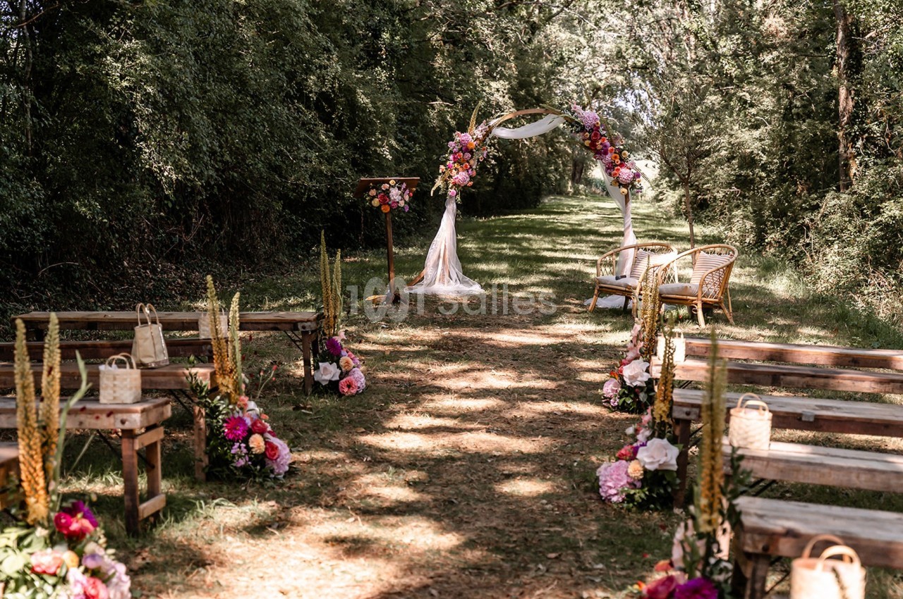 Allée décorée de fleurs menant à une arche nuptiale en extérieur, entourée d'arbres et de bancs en bois.