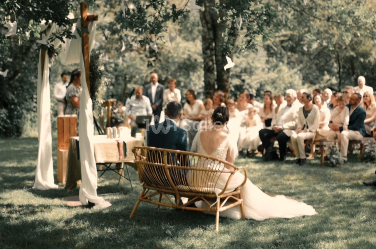 Un couple assis sur un banc en osier lors d'une cérémonie de mariage en plein air, entouré d'invités.