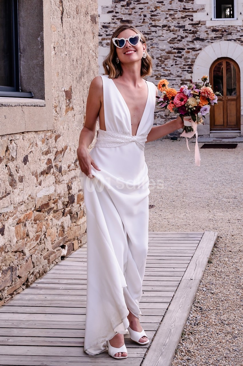 Femme souriante en robe de mariée blanche tenant un bouquet de fleurs colorées devant un bâtiment en pierre.