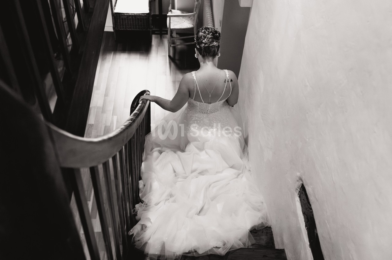 Une femme en robe de mariée descend un escalier en bois dans un intérieur lumineux.
