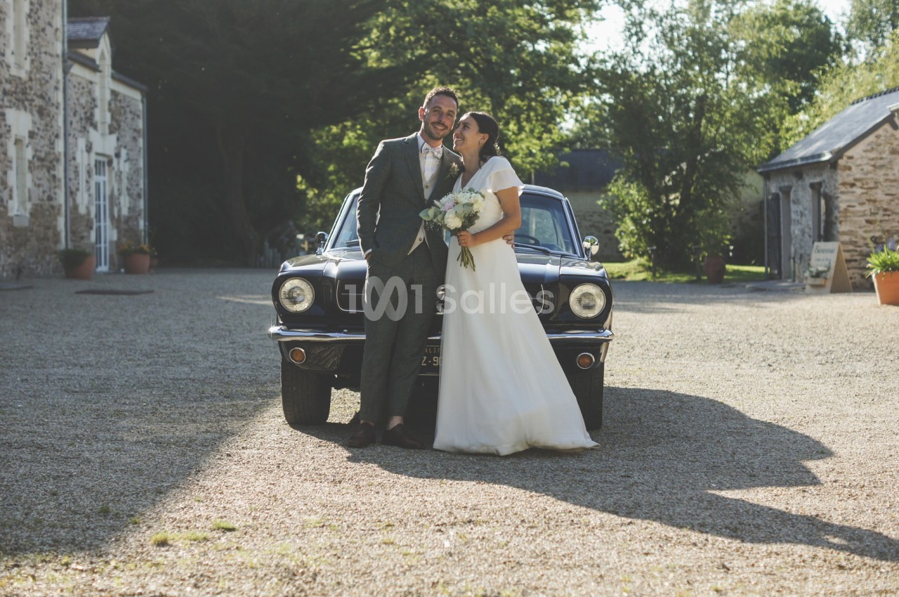 Un couple en tenue de mariage pose devant une voiture ancienne dans une cour ensoleillée bordée de bâtiments en pierre.
