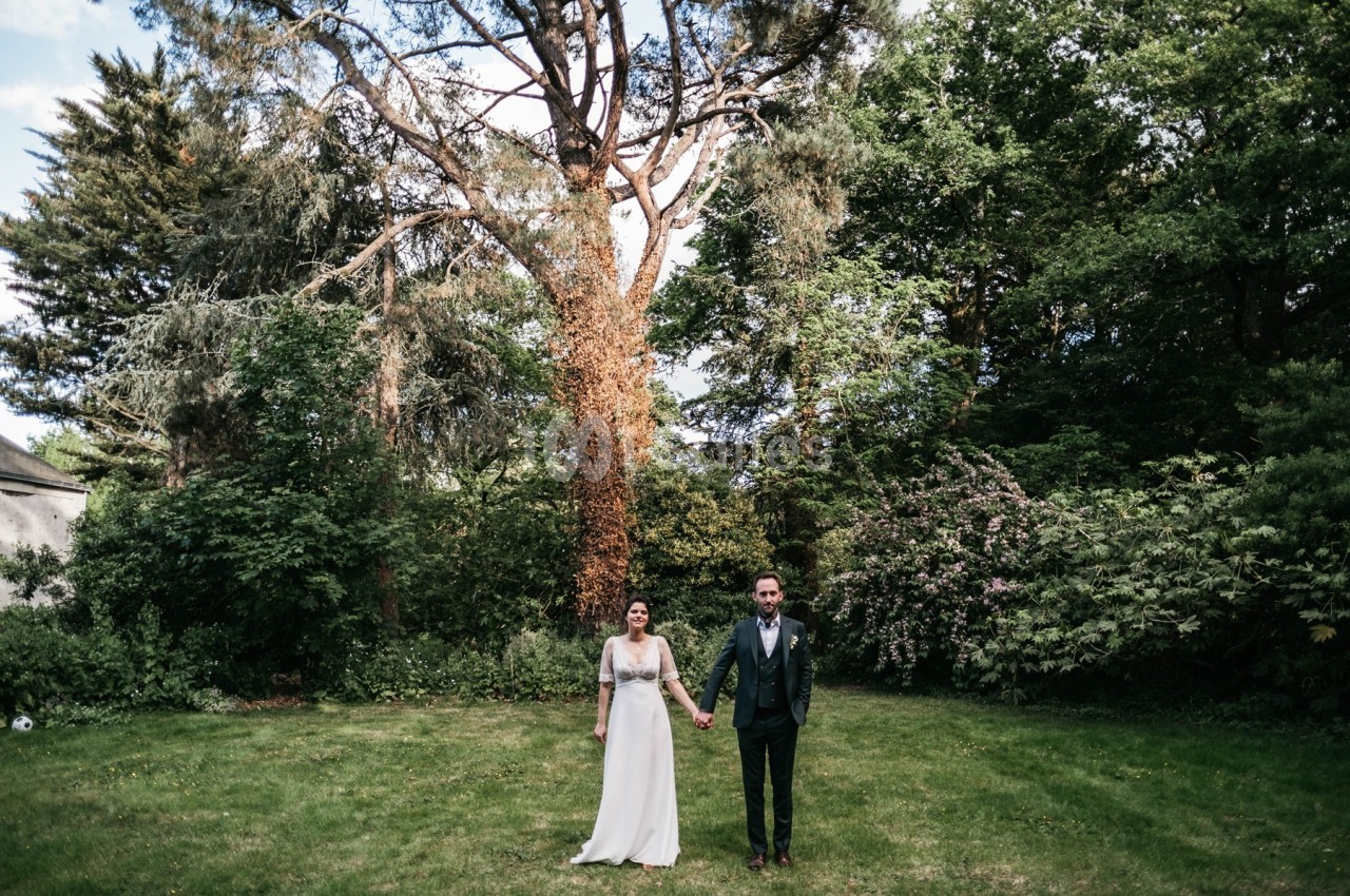 Un couple en tenue de mariage se tient la main dans un jardin verdoyant, entouré d'arbres et de buissons.