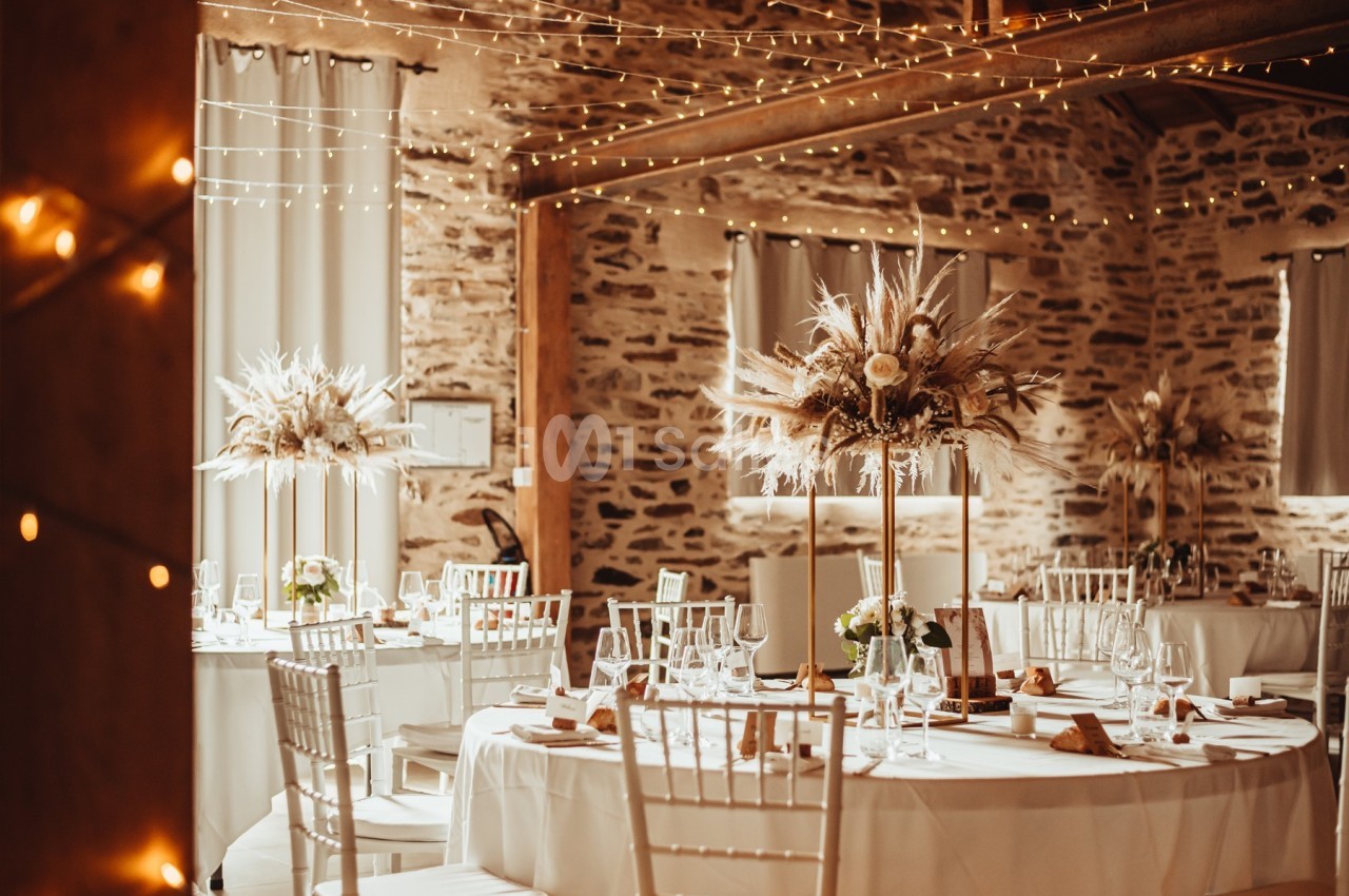 Salle de réception décorée avec des tables rondes, des chaises blanches et des centres de table floraux sous des guirlandes…