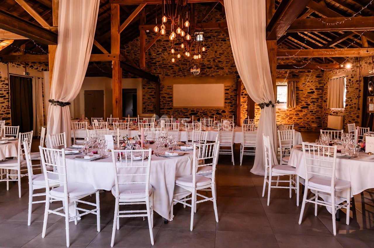 Salle de réception rustique avec murs en pierre, tables rondes dressées et chaises blanches sous un éclairage chaleureux.