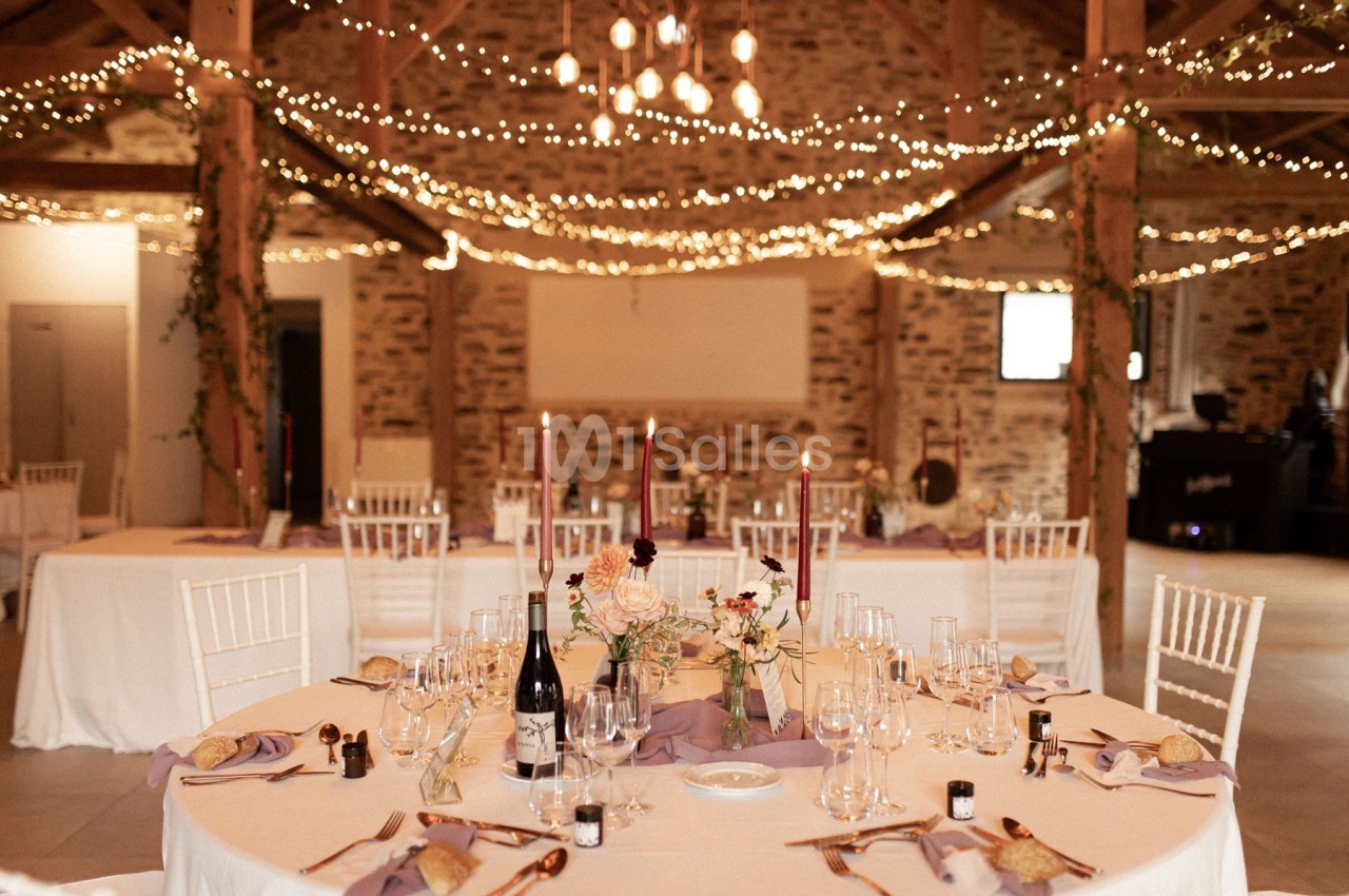 Salle de réception décorée avec des guirlandes lumineuses, tables dressées avec nappes blanches et centre de table floral.