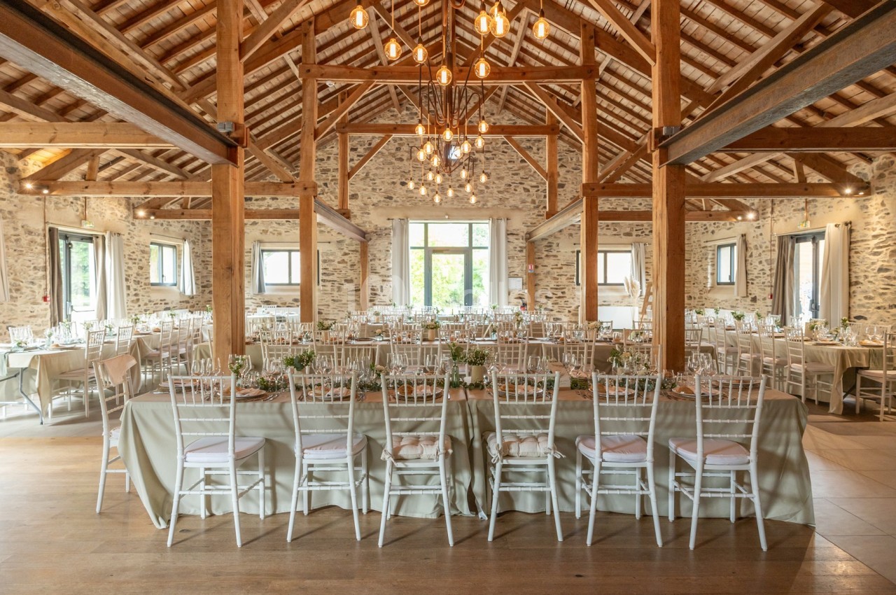 Salle de réception lumineuse avec charpente en bois, tables décorées et chaises blanches alignées.