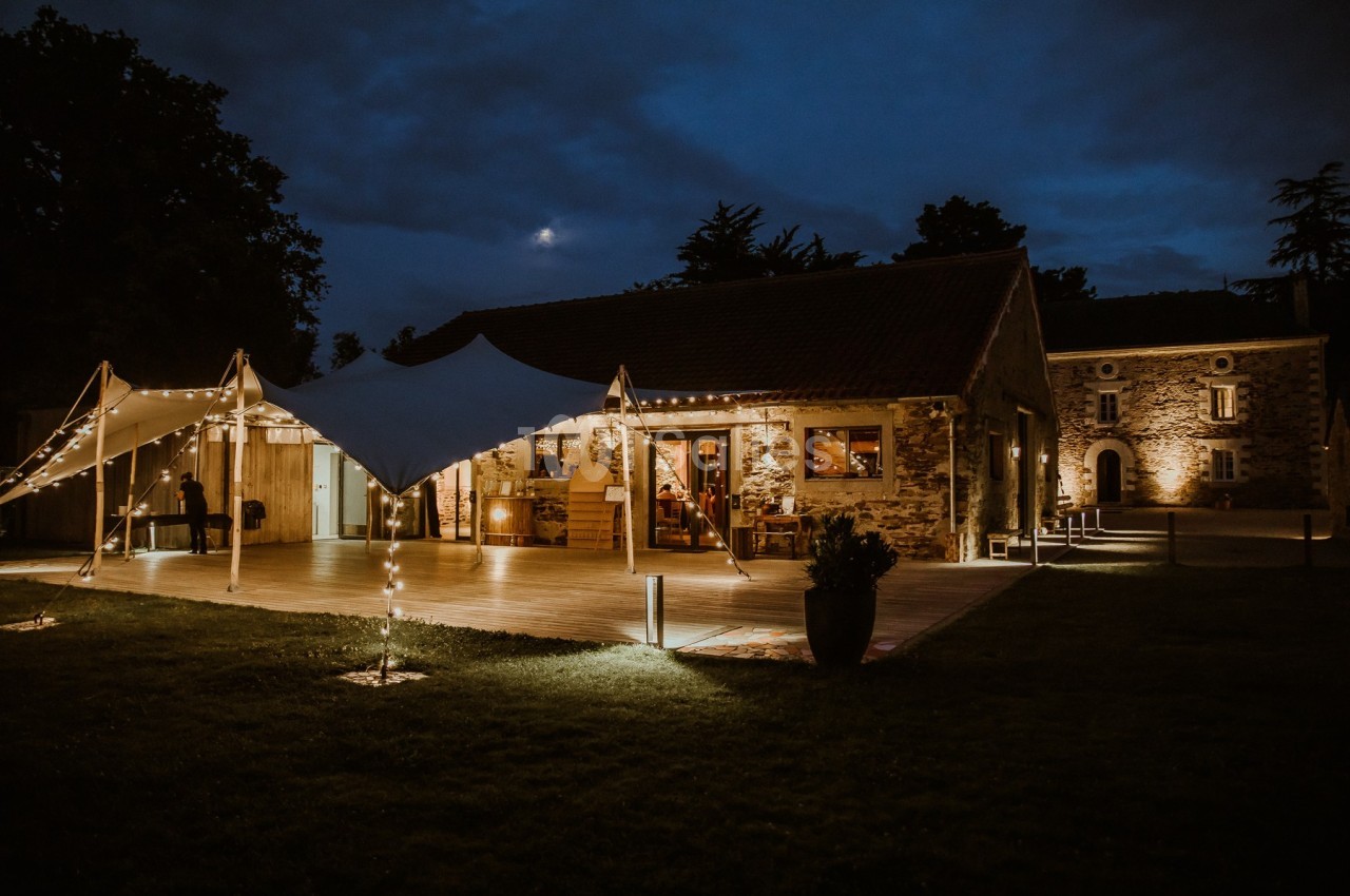 Bâtiment en pierre éclairé de nuit avec une tente lumineuse installée sur une terrasse en bois.