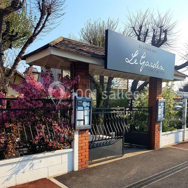 Entrée d'un restaurant avec une enseigne ’Le Garden’, entourée de fleurs roses et d'arbres, sous un ciel clair.