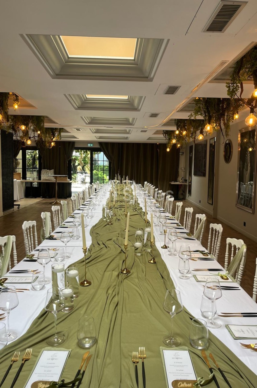 Salle intérieure dressée en banquet avec table longue, nappe verte, chaises blanches et décoration florale suspendue.