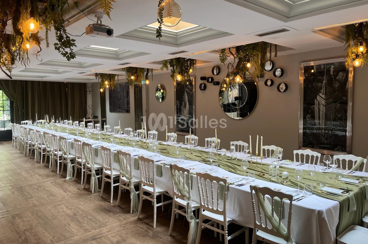 Salle intérieure dressée avec long banquet blanc, chaises chiavari blanches, décoration florale verte suspendue et éclairage doré.