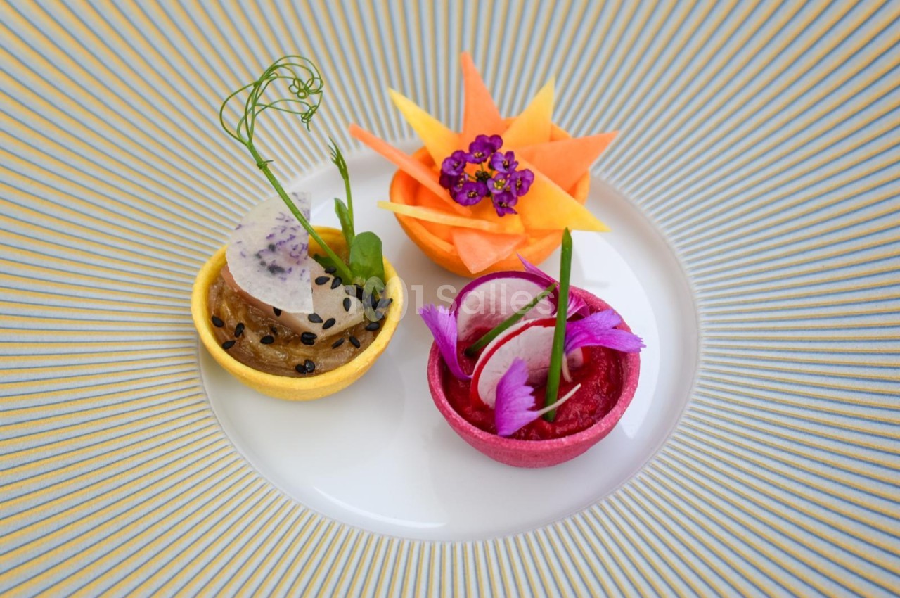 Assiette blanche avec trois amuse-bouches colorés - mousse en coque jaune, betterave en coque rose, fleur orange et jaune - sur fond rayé bleu et or.