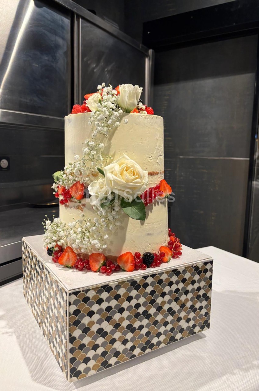 Gâteau de mariage trois étages blanc crème, orné de roses blanches, fleurs rouges et petites fleurs blanches, sur socle motifs géométriques.
