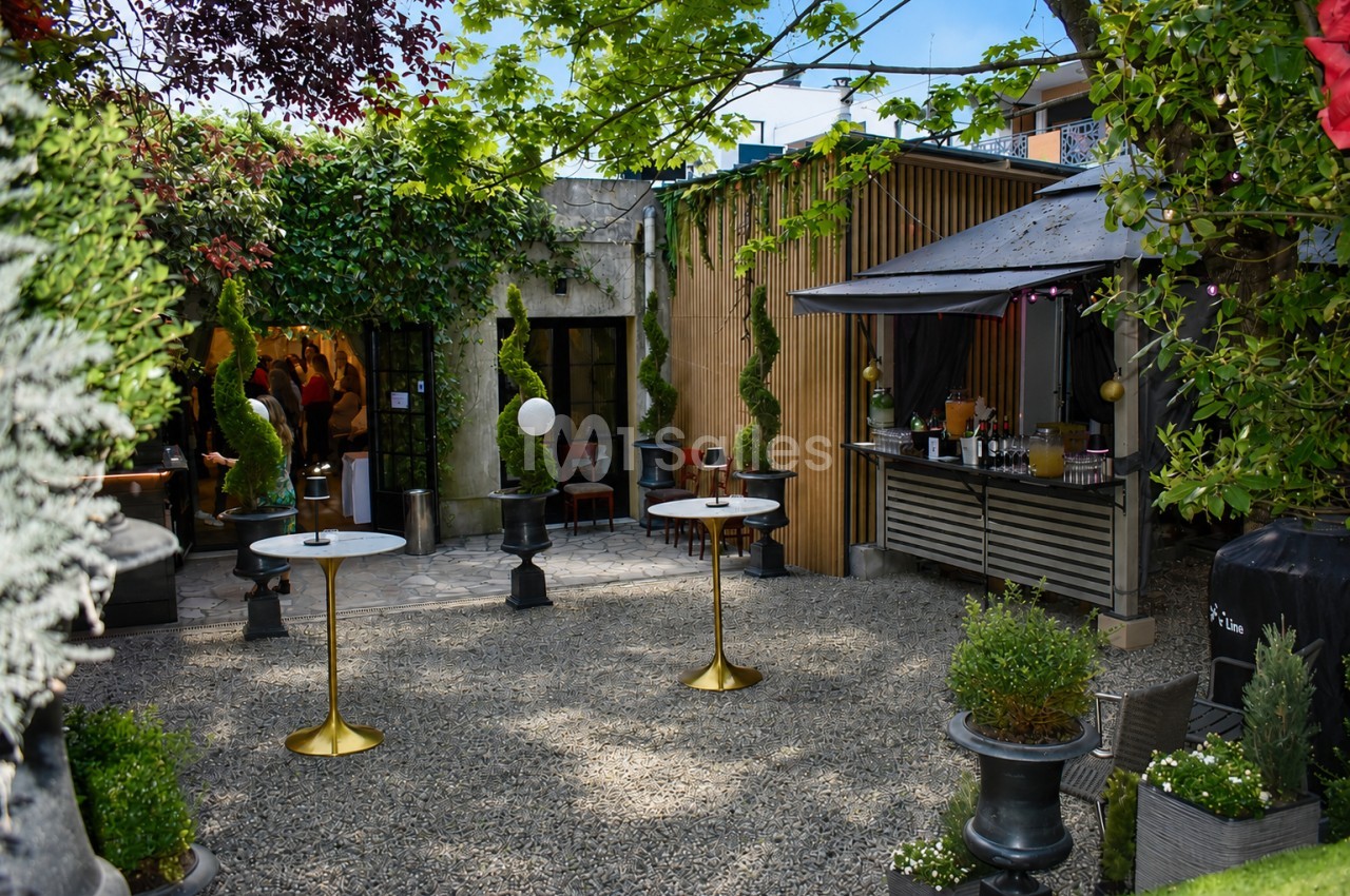 Cour intérieure paysagée avec tables hautes, terrasse couverte et façades en bois clair.
