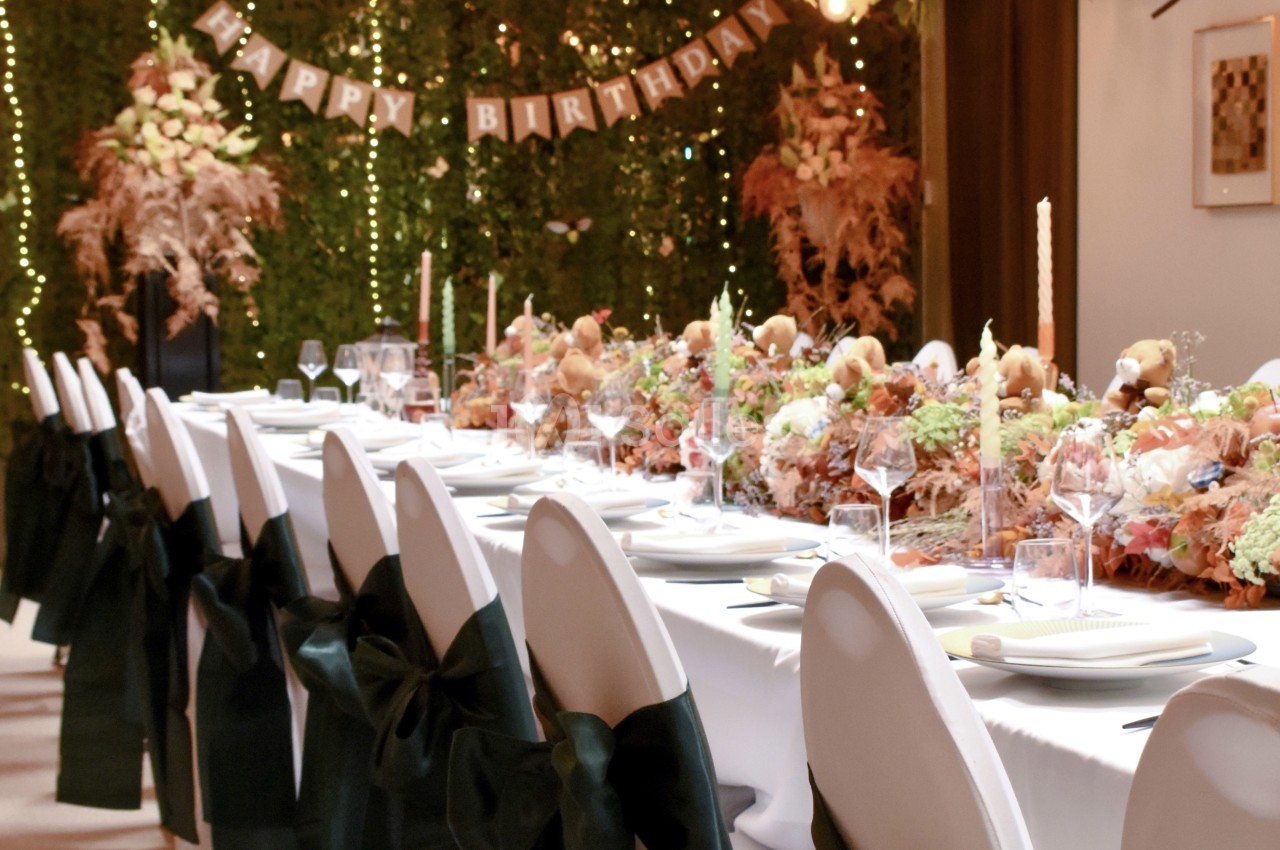 Salle dressée en banquet avec table longue, chaises blanches et noires, bouquets floraux roses et blancs, guirlande lumineuse et feuillage en arrière-plan.