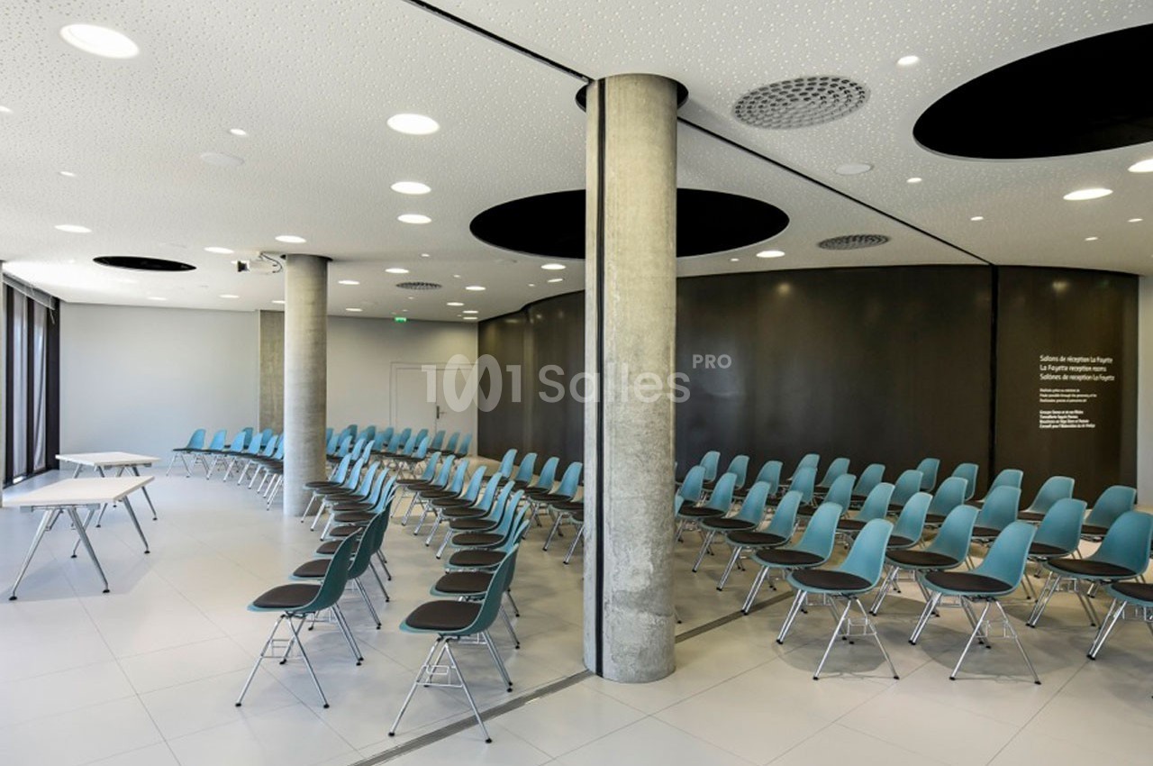 Salle de conférence moderne avec des rangées de chaises bleues, murs sombres et colonnes en béton apparent.