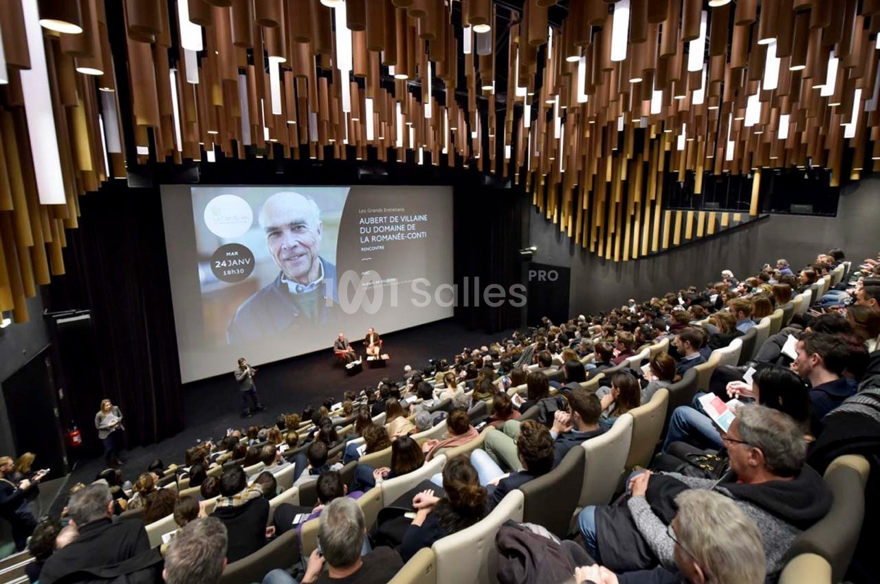 Salle de conférence remplie, avec un écran projetant une image et des intervenants sur scène.