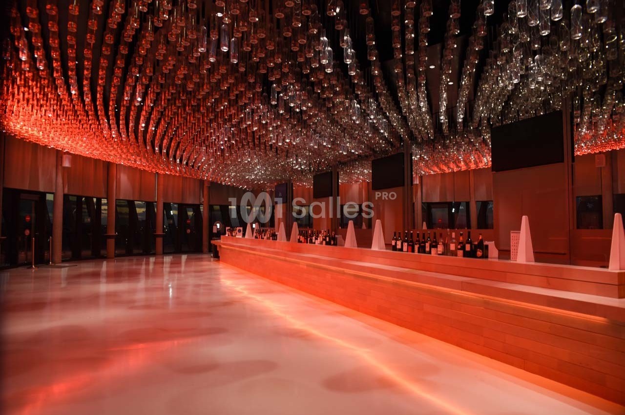 Plafond décoré de bouteilles suspendues, éclairage rouge, bar en bois long et moderne dans une grande salle.
