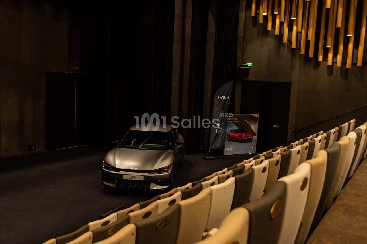 Une voiture grise exposée sur une scène dans une salle sombre avec des rangées de sièges et un panneau promotionnel.