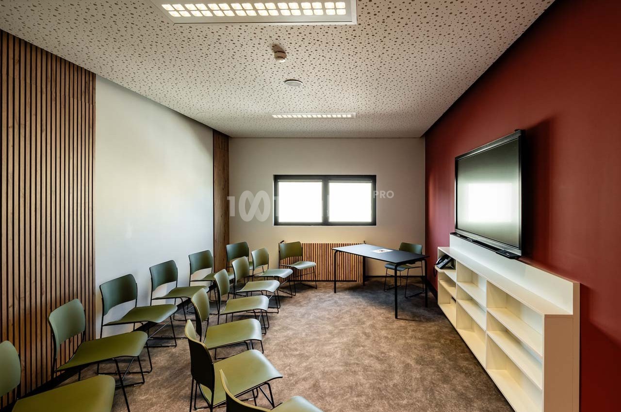 Salle de réunion avec chaises disposées en arc de cercle, table, étagère blanche et écran mural.