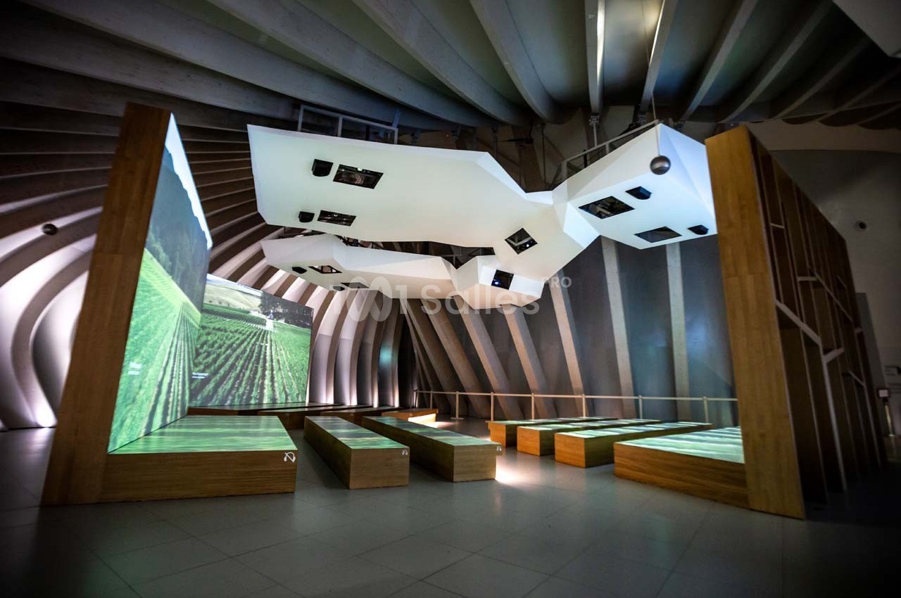 Salle moderne avec bancs en bois, écrans suspendus et projections lumineuses sur le sol, entourée d'arches métalliques.