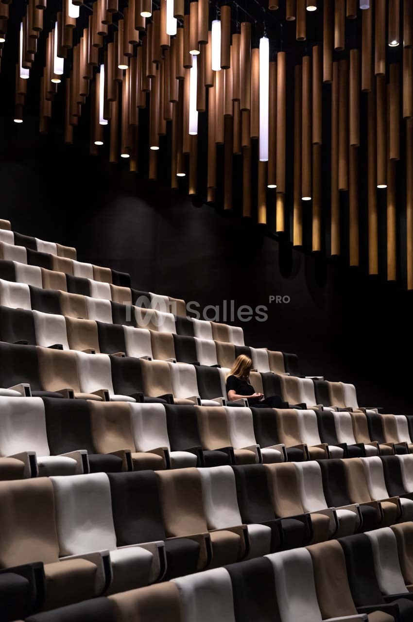 Une femme assise seule dans un auditorium aux sièges beige et noir, éclairé par des suspensions modernes.