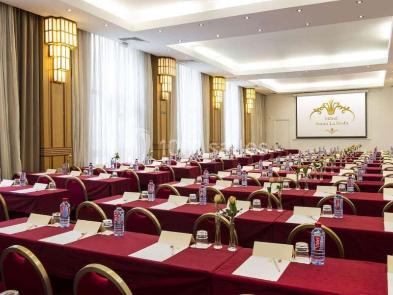 Salle de conférence lumineuse avec des rangées de tables rouges, chaises assorties et un écran de projection au fond.