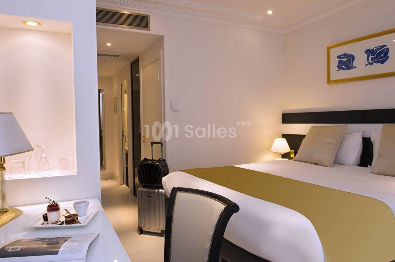 Chambre d'hôtel lumineuse avec lit double, valise posée près de la porte et bureau avec tasse et dessert.