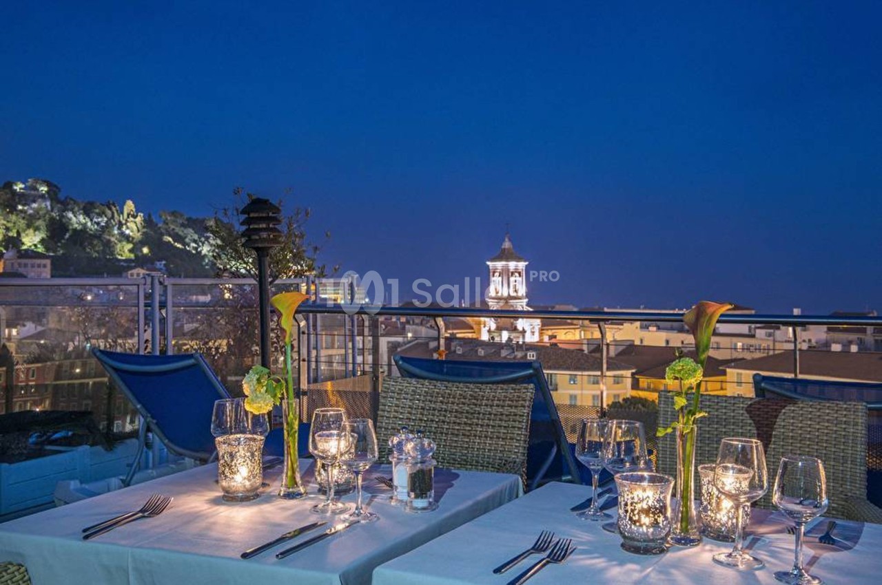 Tables dressées avec chandelles et fleurs sur une terrasse offrant une vue nocturne sur des bâtiments éclairés.