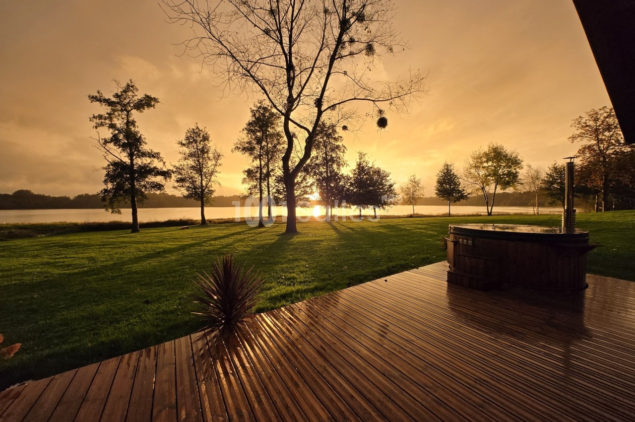 Coucher de soleil sur un lac, avec des arbres en contre-jour et une terrasse en bois au premier plan.
