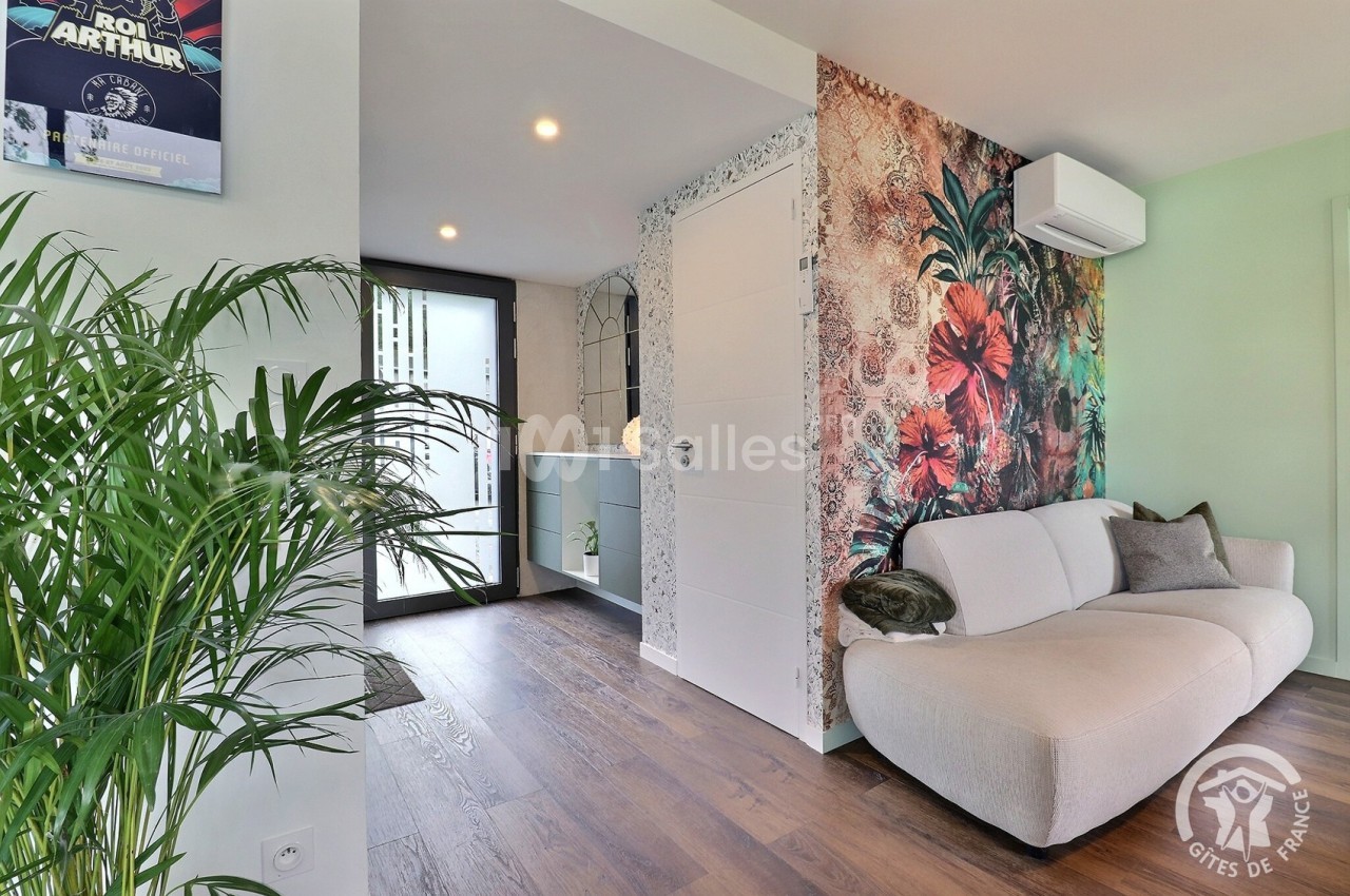 Salon lumineux avec canapé beige, mur décoré de motifs floraux, sol en parquet et vue sur une entrée moderne.