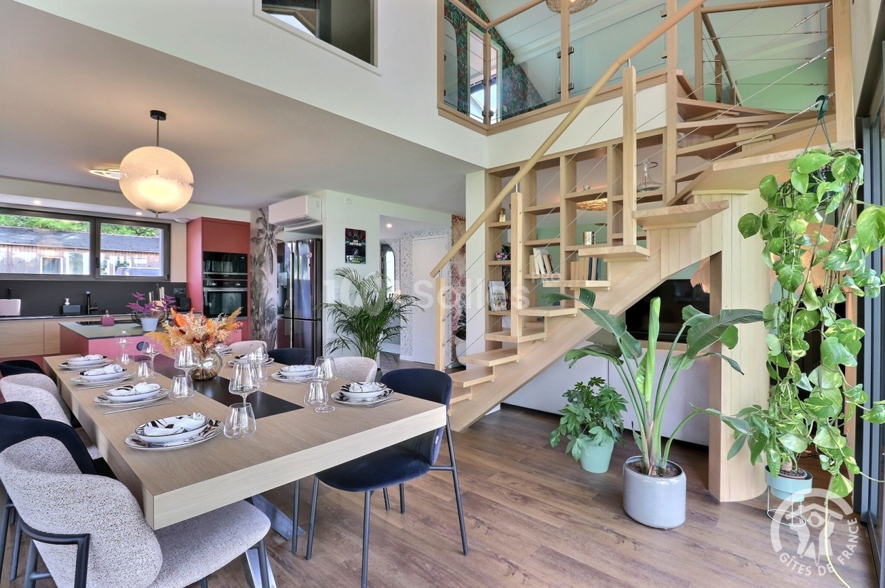 Salle à manger moderne avec table dressée, cuisine ouverte en arrière-plan et escalier en bois menant à l'étage.