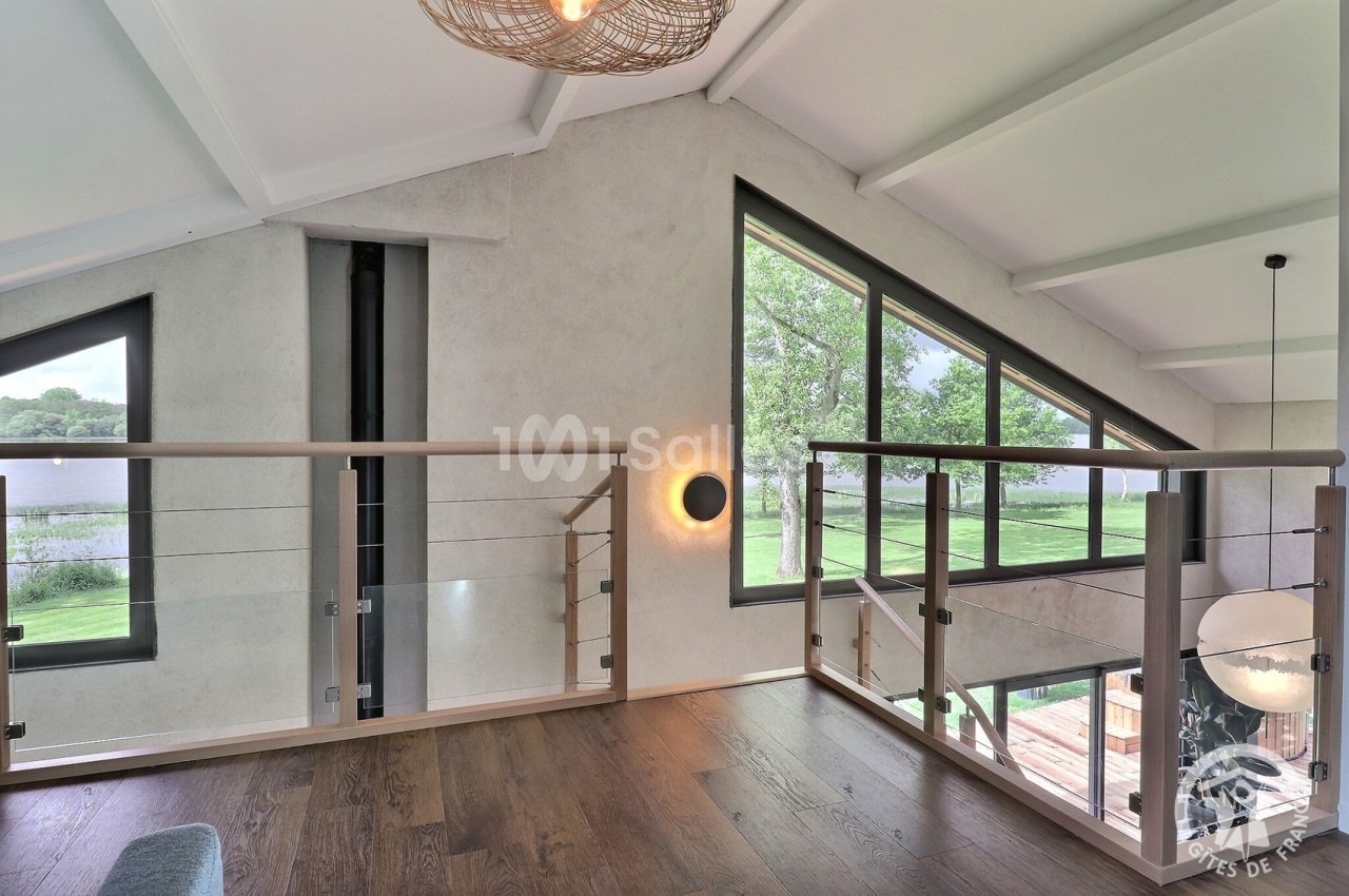 Mezzanine lumineuse avec parquet en bois, garde-corps en verre et vue sur un paysage verdoyant à travers de grandes fenêtres.