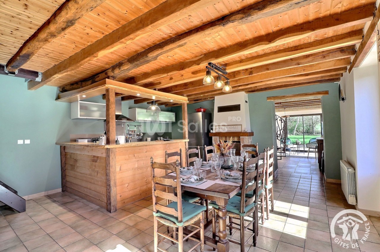 Salle à manger lumineuse avec table en bois, cuisine ouverte et poutres apparentes, donnant sur un jardin.