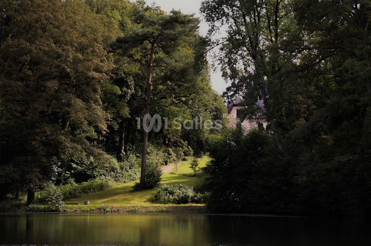 Vue d'un parc verdoyant avec un plan d'eau au premier plan et un bâtiment partiellement visible entre les arbres.