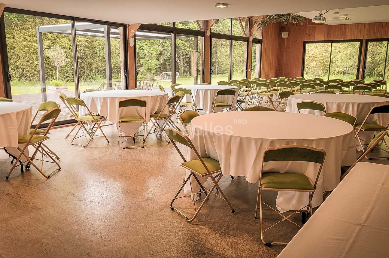 Salle lumineuse avec grandes baies vitrées, tables rondes dressées de nappes blanches et chaises pliantes vertes.