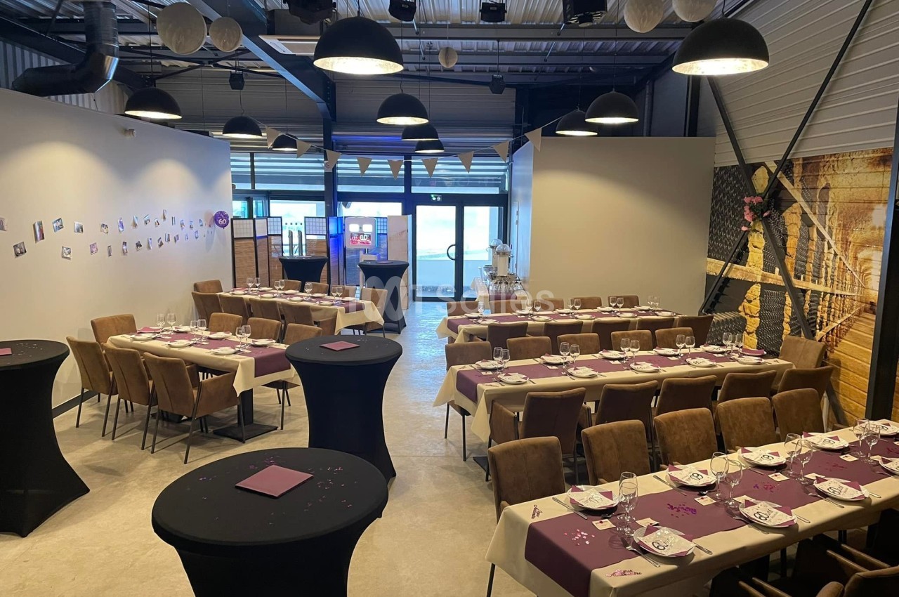 Salle décorée pour un événement avec tables dressées, nappes violettes, chaises rembourrées et éclairage suspendu.