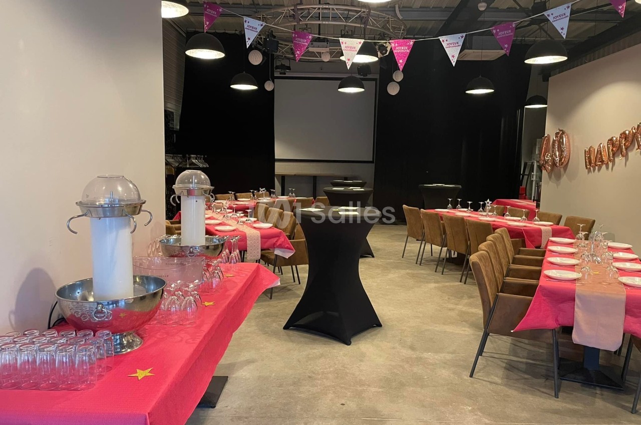 Salle décorée pour un événement avec tables dressées, nappes roses, guirlandes triangulaires et projecteur en fond.