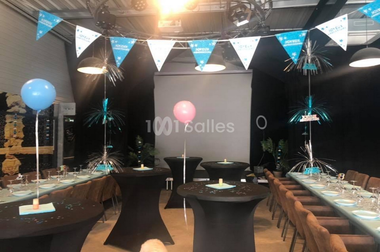 Salle décorée pour une fête avec des ballons, guirlandes et tables dressées sous un éclairage tamisé.