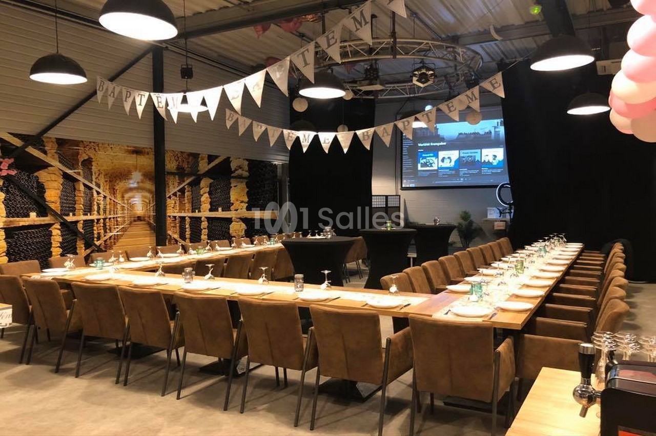 Salle décorée pour un événement avec longues tables dressées, guirlandes suspendues et écran en fond.