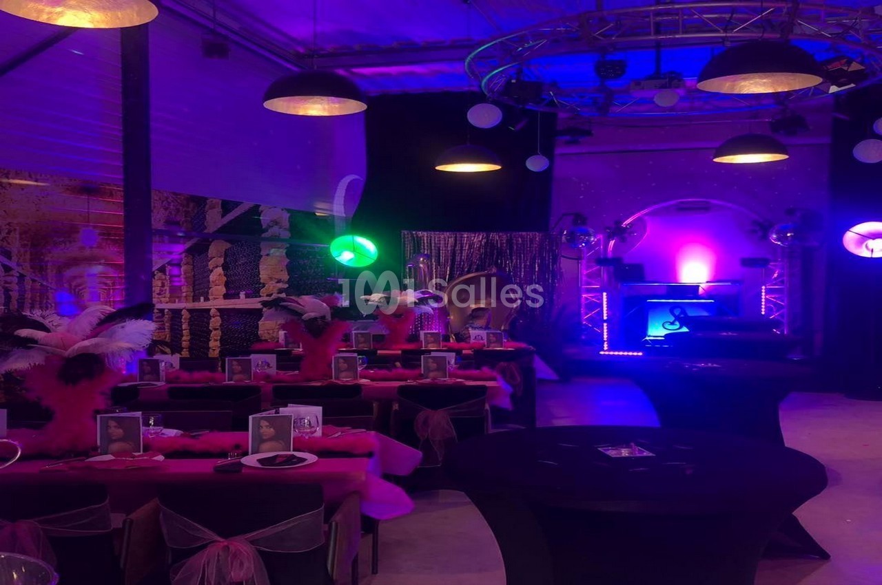 Salle décorée pour un événement festif avec tables ornées de plumes, éclairage coloré et espace DJ illuminé.