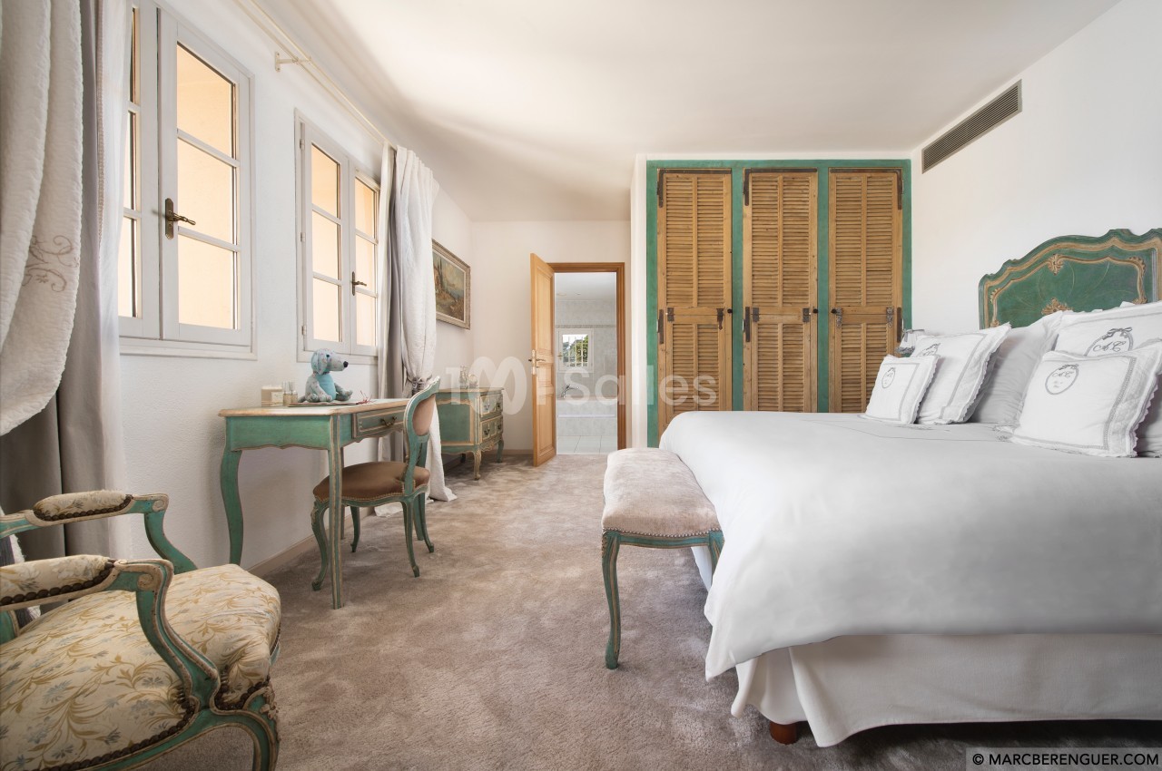 Chambre lumineuse avec lit double, mobilier ancien, bureau et armoire en bois, fenêtres donnant sur l'extérieur.
