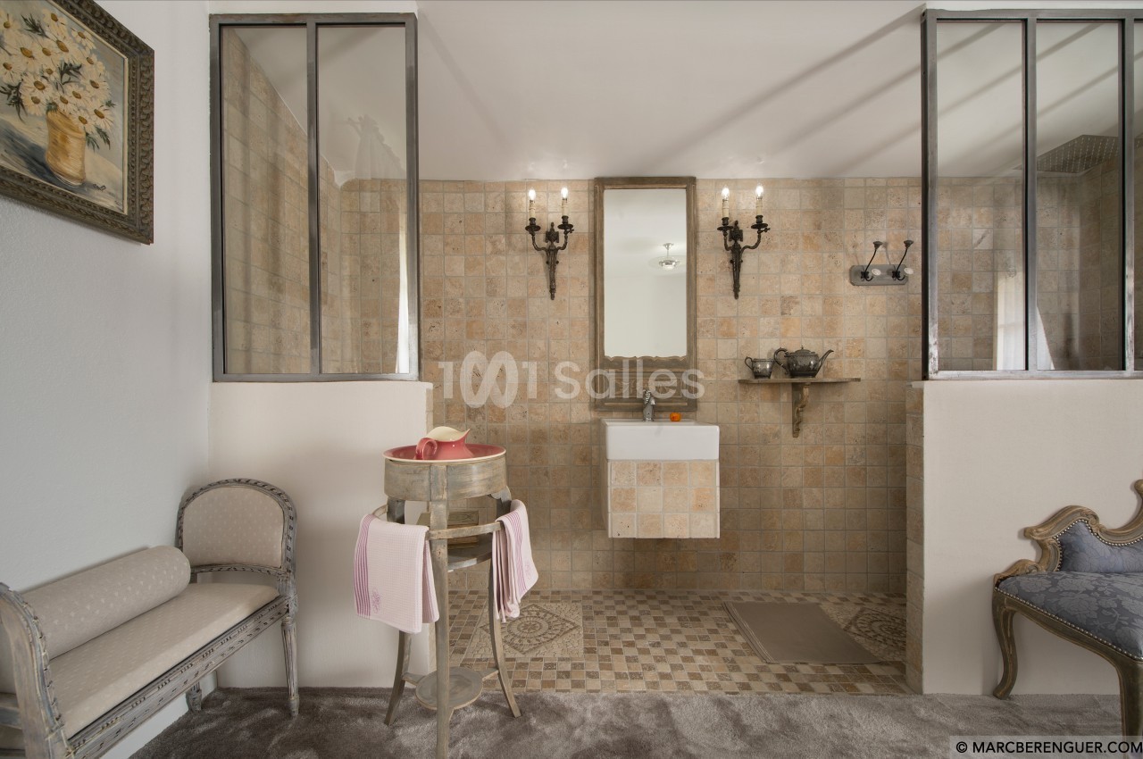 Salle de bain rustique avec carrelage beige, lavabo suspendu, miroir encadré, et mobilier vintage en bois et tissu.