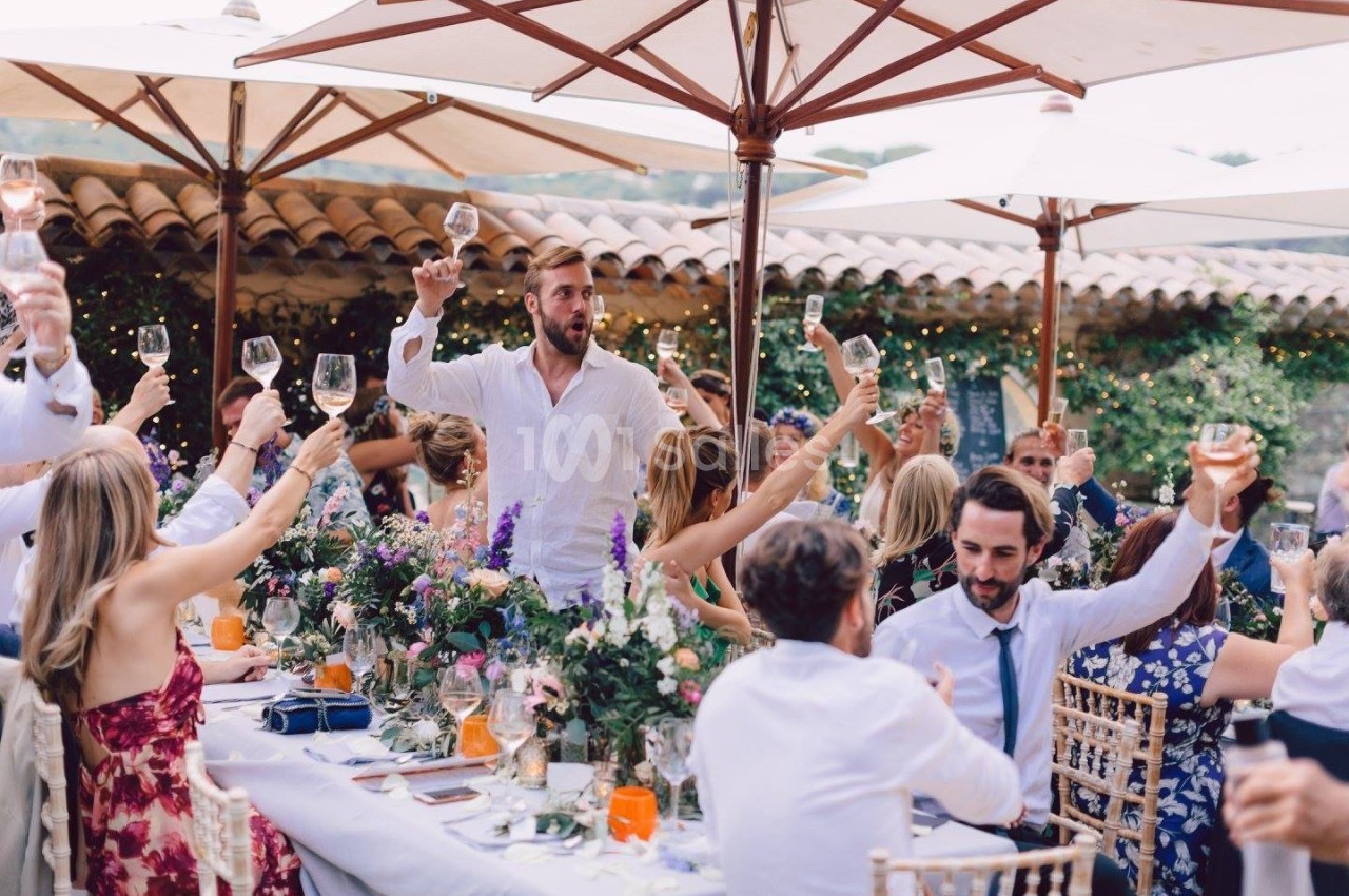 Des convives lèvent leurs verres autour d'une grande table décorée, sous des parasols, dans un cadre extérieur festif.