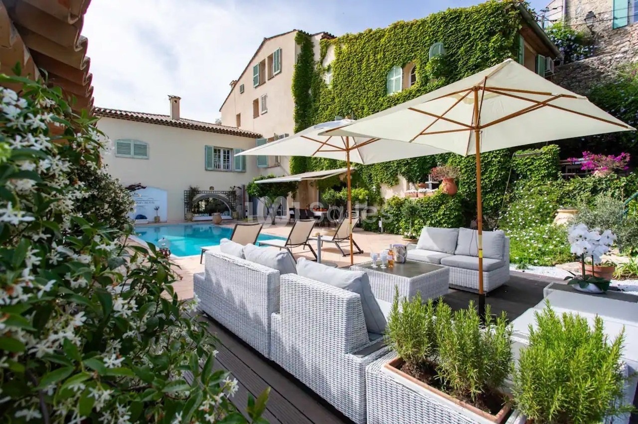 Terrasse avec mobilier en rotin, parasols et piscine, entourée de végétation et d'une maison couverte de lierre.