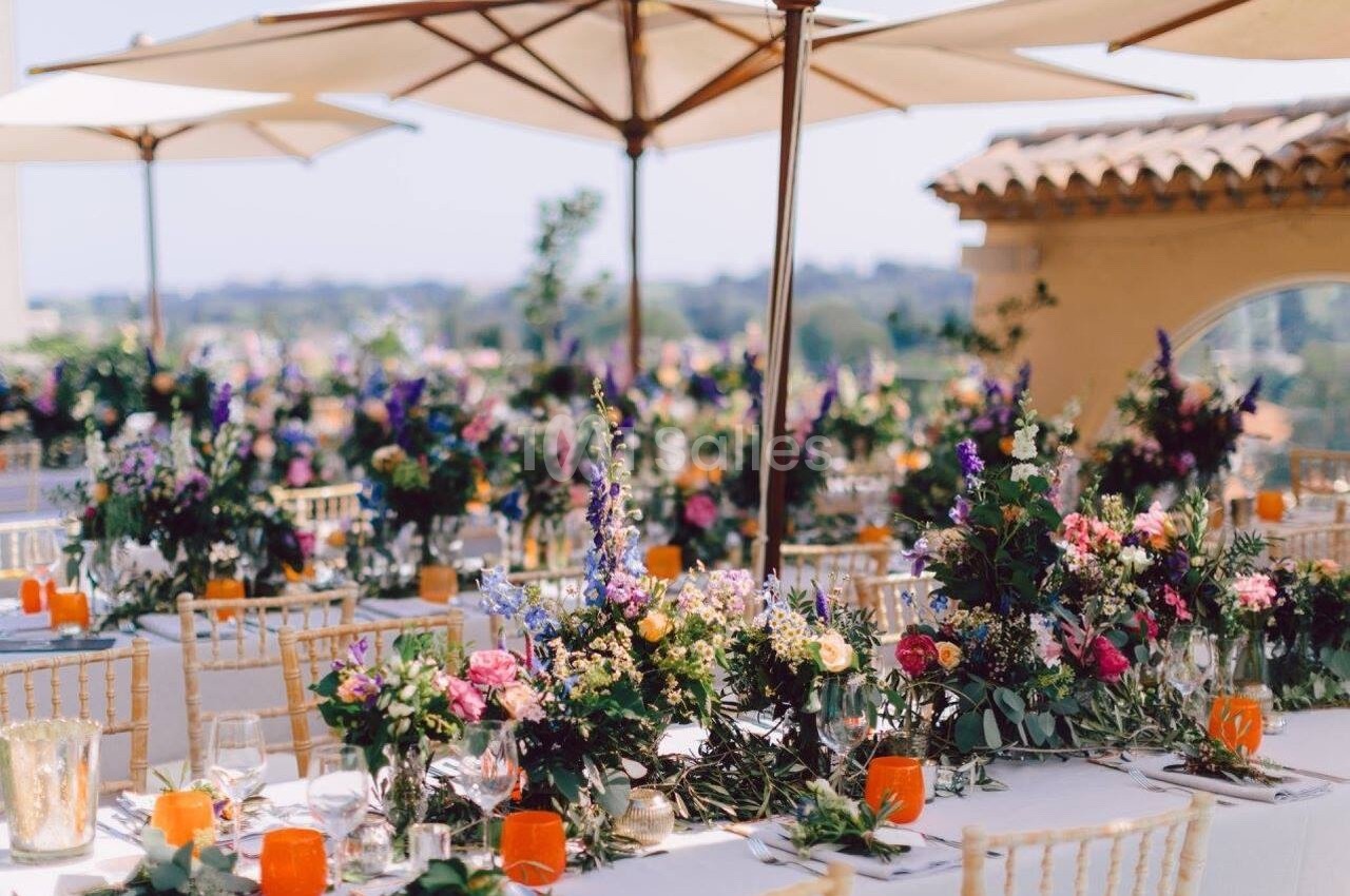 Tables décorées de fleurs colorées et de bougies oranges sous des parasols, en extérieur par temps ensoleillé.