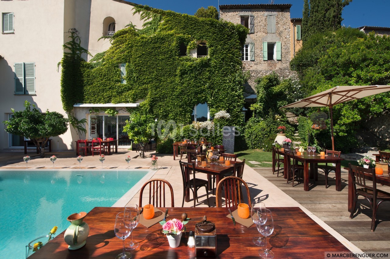Terrasse avec tables dressées près d'une piscine, entourée de bâtiments couverts de végétation et de verdure.