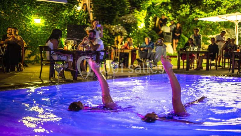 Deux personnes exécutent une chorégraphie synchronisée dans une piscine éclairée, entourée de spectateurs en soirée.
