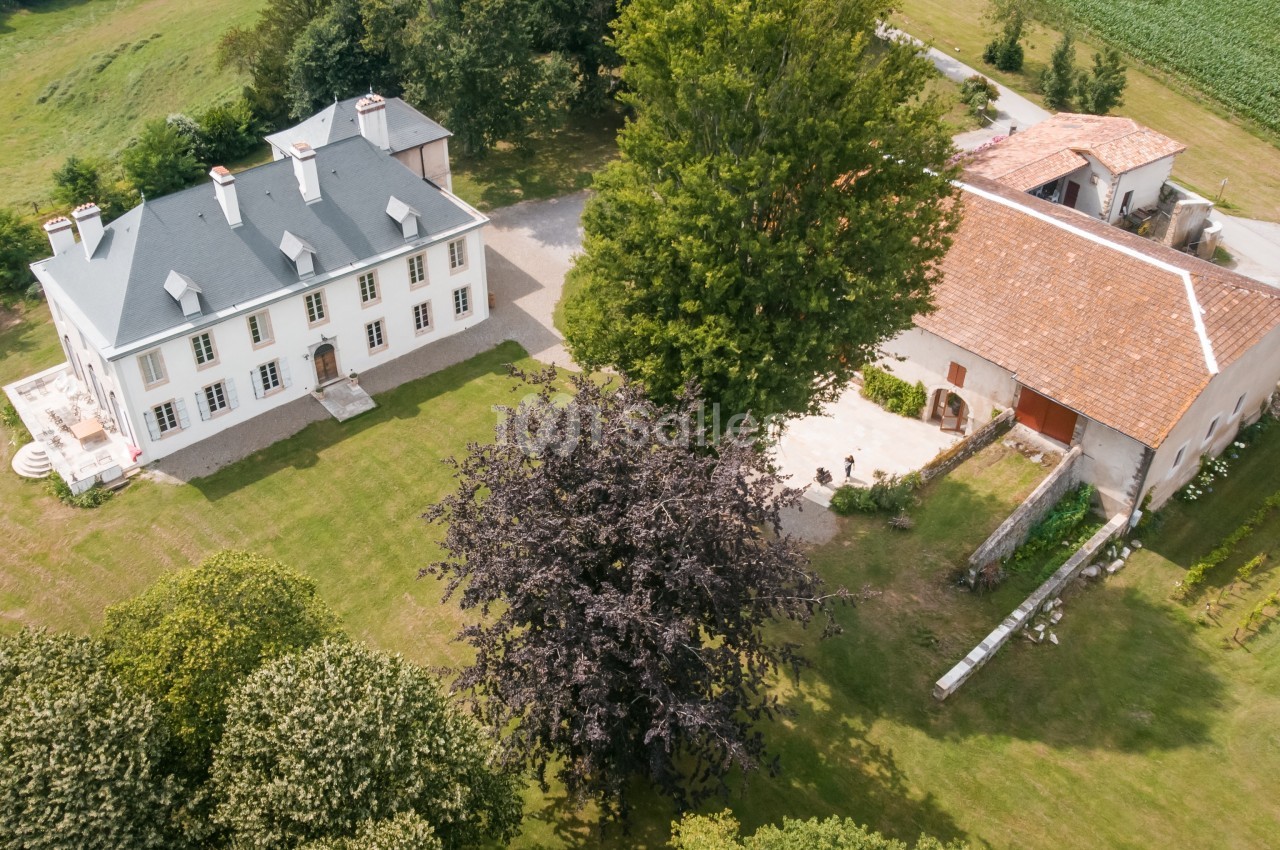 Vue aérienne d'une maison de campagne avec un toit gris, une grange attenante et un grand jardin arboré.