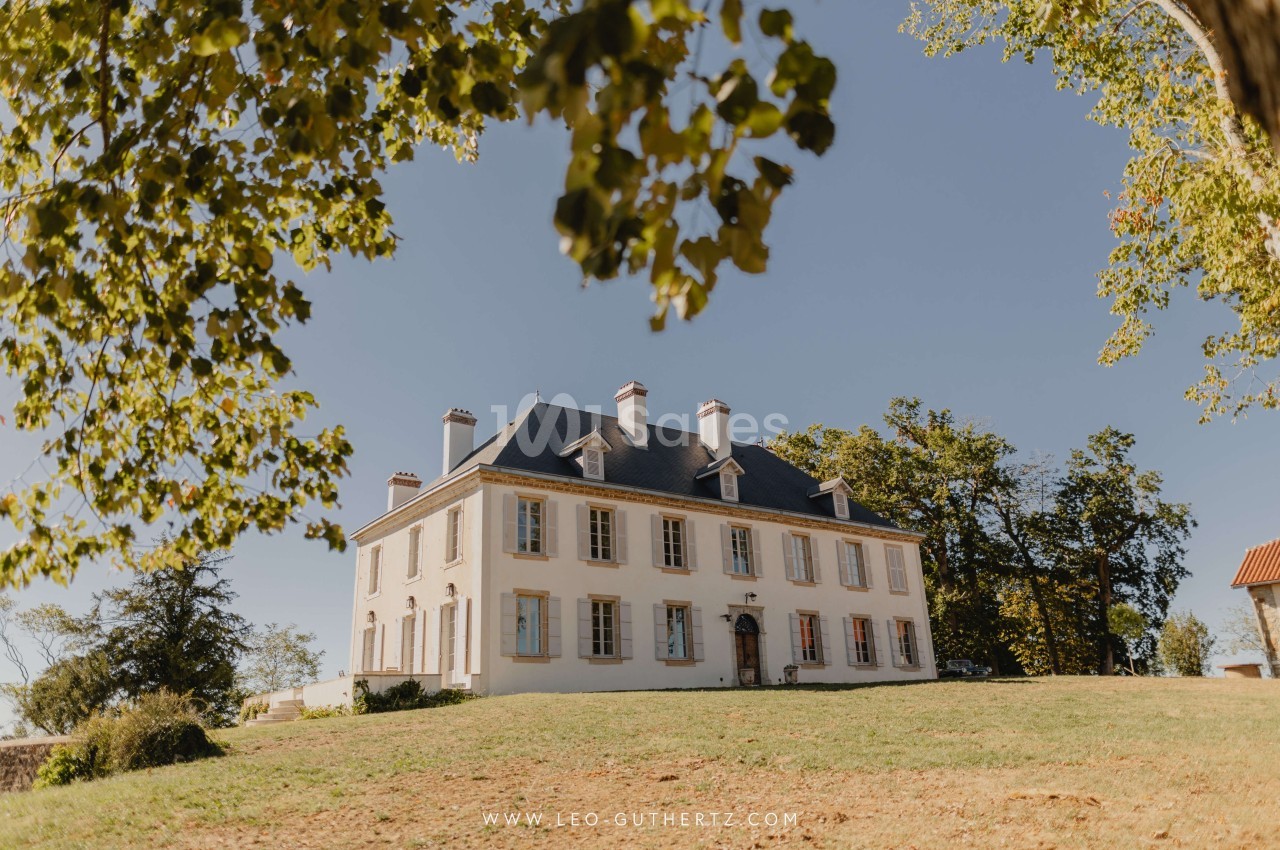 Manoir blanc à deux étages avec toit en ardoise, entouré d'arbres et d'une pelouse sous un ciel dégagé.